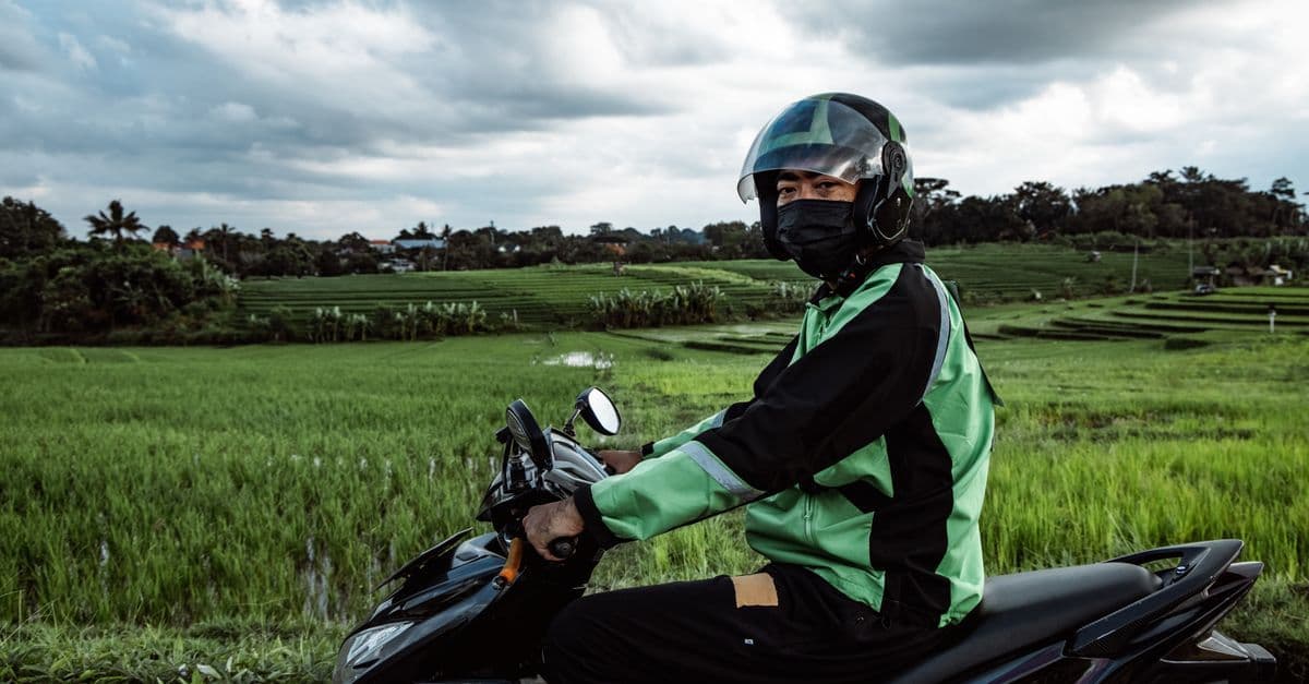 A masked motorcyclist in a helmet sits on a scooter beside lush rice fields in Indonesia.