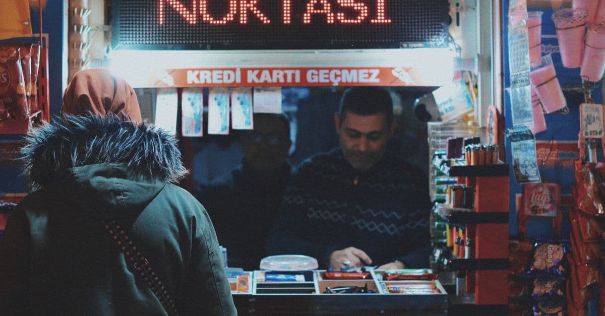 Person mit Kapuzenmantel steht vor einem kleinen Kiosk, Verkäufer zählt Geld hinter einem voll bestückten Tresen.