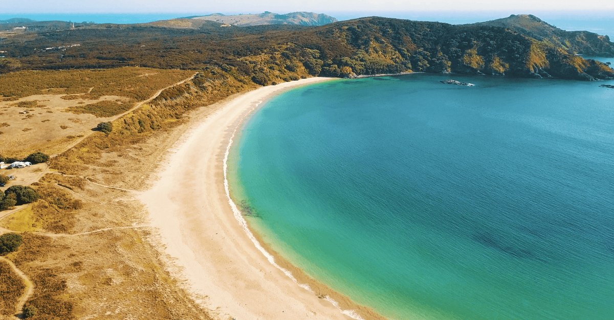 A sweeping crescent of pale sand borders a turquoise bay beneath dry hills and distant mountains.