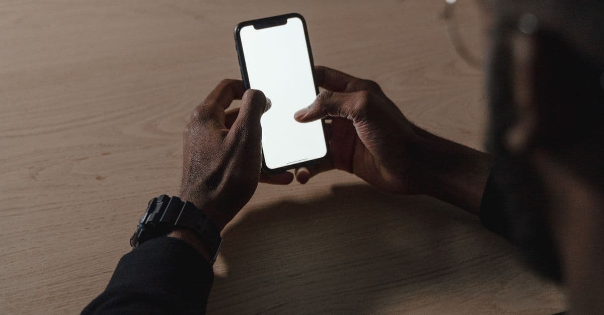A person holding a smartphone with a blank white screen over a wooden table, seen from above.
