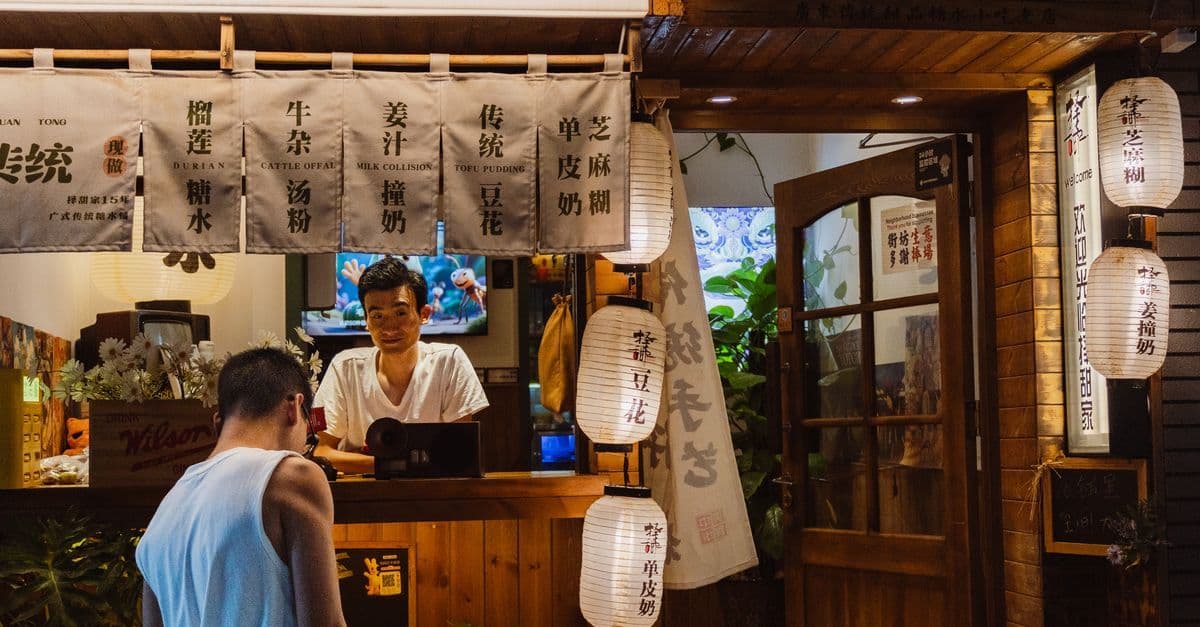 A customer talks with a worker at a warmly lit Chinese dessert shop under hanging menu banners and paper lanterns.