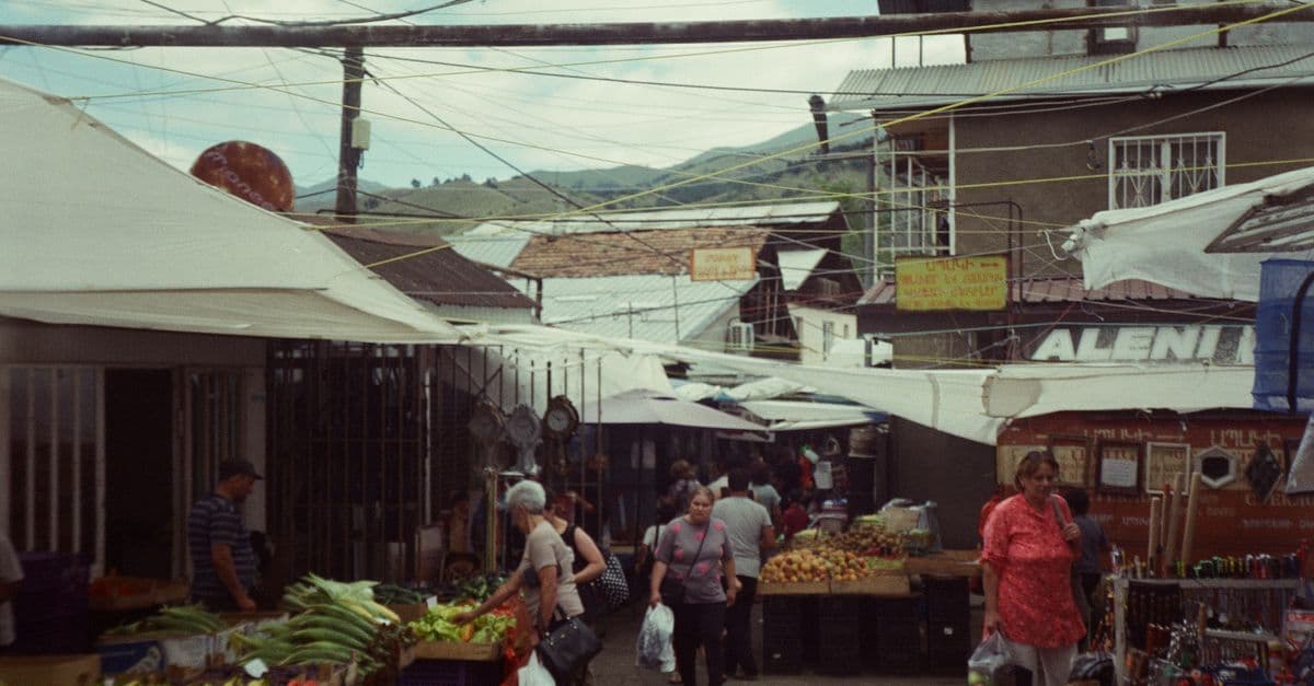 Shoppers walk through a busy street market lined with produce stalls and overhead wires.