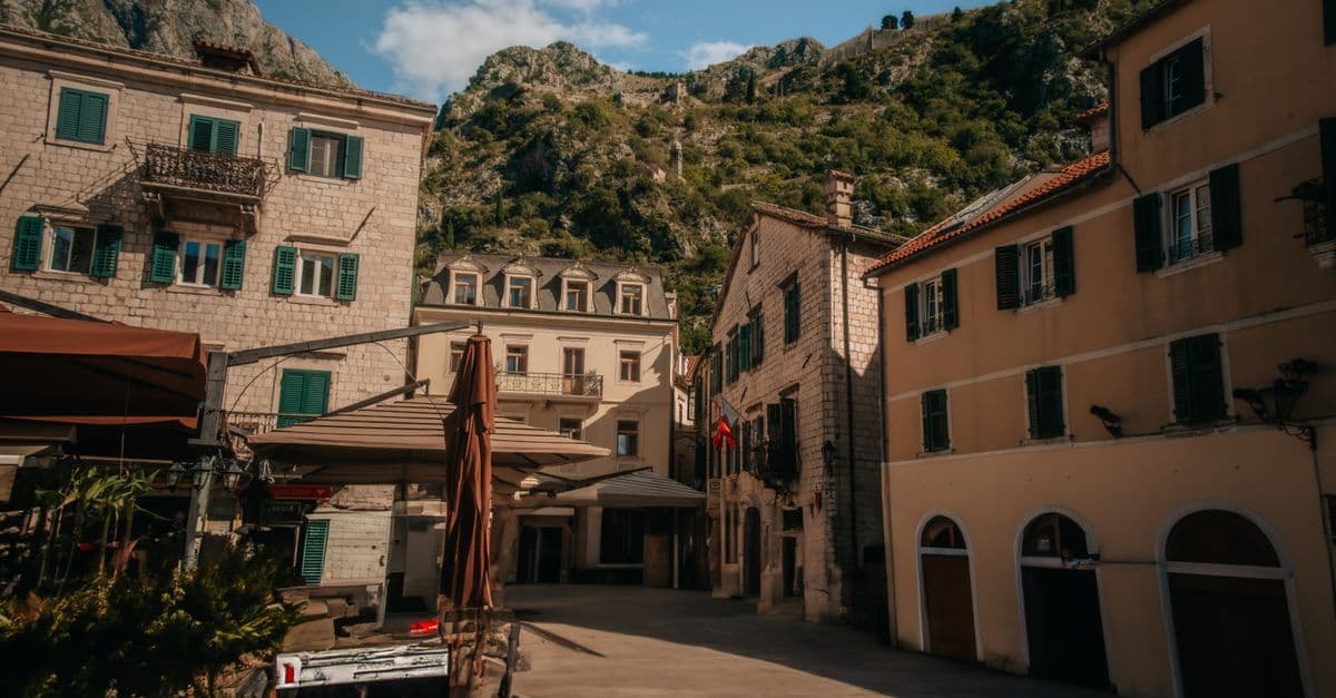 Sunlit Mediterranean town square with stone buildings, green shutters, outdoor cafe umbrellas, and rocky hills rising behind.