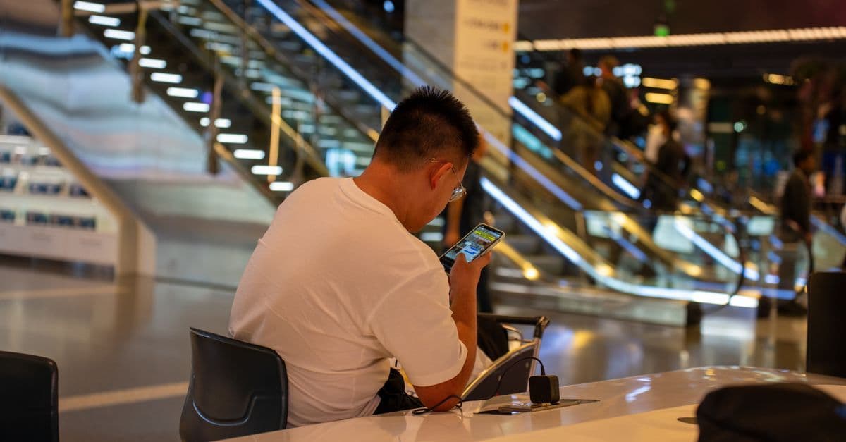 Erwachsener nutzt Smartphone im modernen Terminal des Hamad International Airport in Doha, belebte Szene mit Rolltreppe.