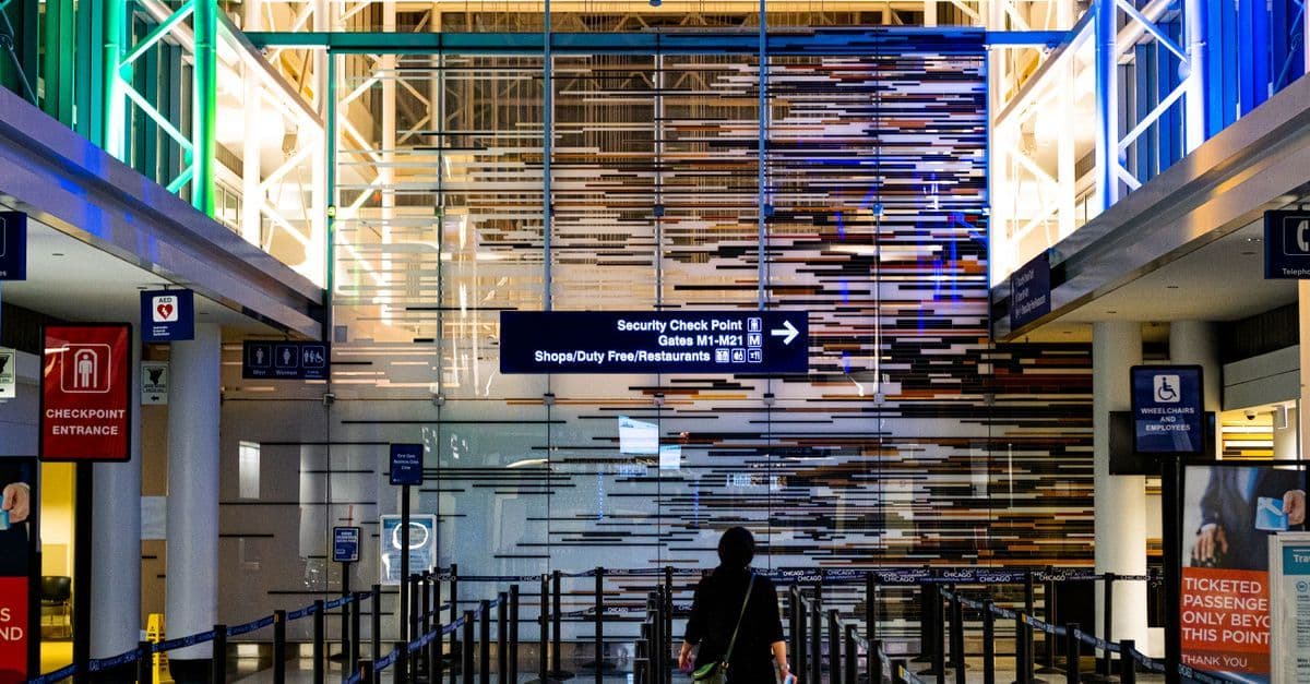 Pasajero frente a un control de seguridad en un aeropuerto, con letreros que indican las puertas de embarque.