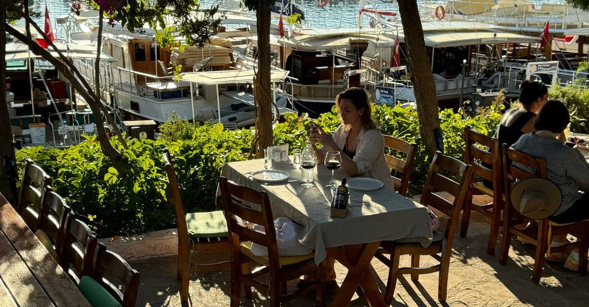 Eine Frau schaut an einem Restauranttisch am Hafen von Kaş auf ihr Smartphone, dahinter liegen Ausflugsboote.