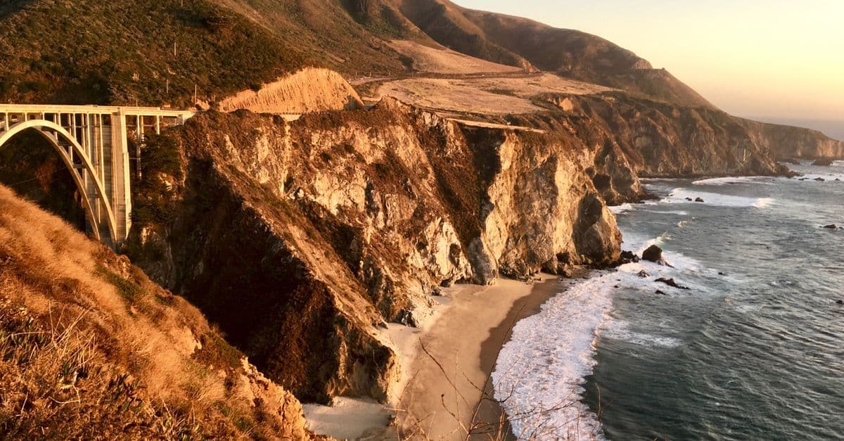 Bixby Creek Bridge och klippkusten i Kalifornien i kvällssol, med vågor som slår in mot en strand.