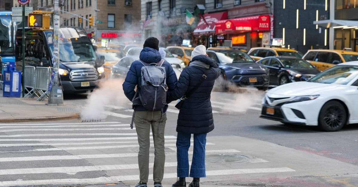 Due viaggiatori con lo zaino aspettano di attraversare una strada di New York, tra taxi gialli e vapore.