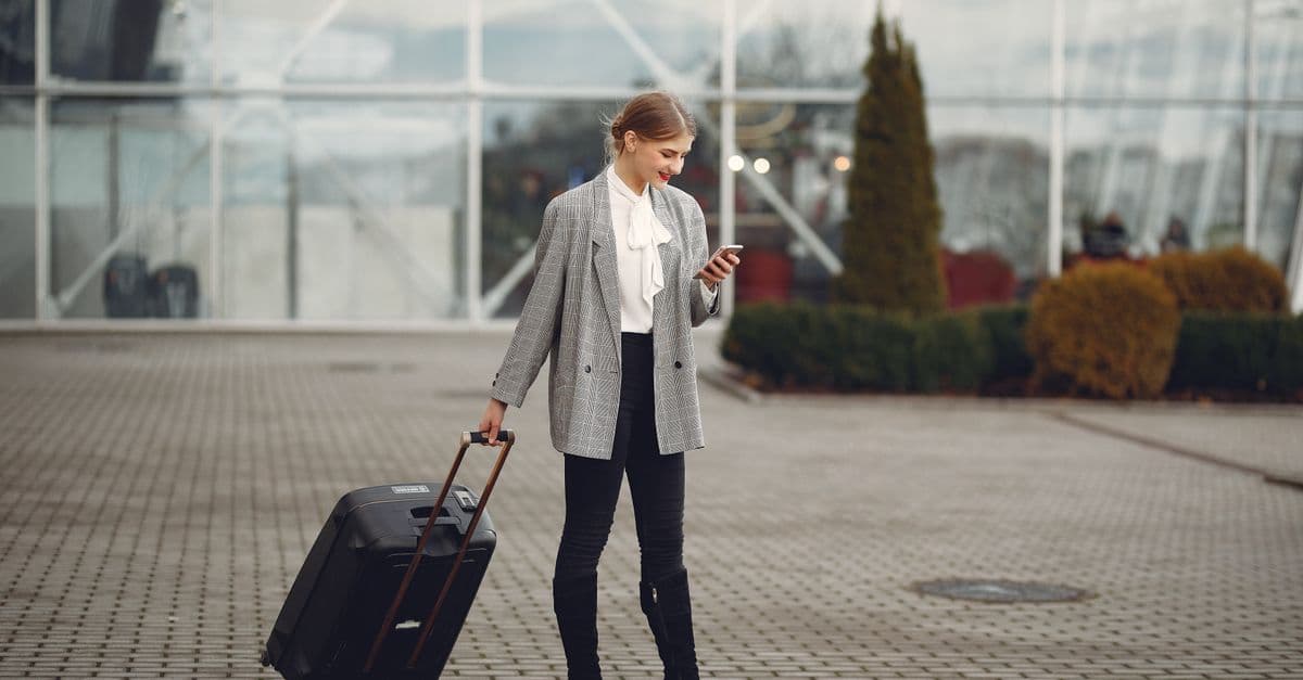 Young woman pulls a wheeled suitcase and looks at her phone outside an airport terminal.