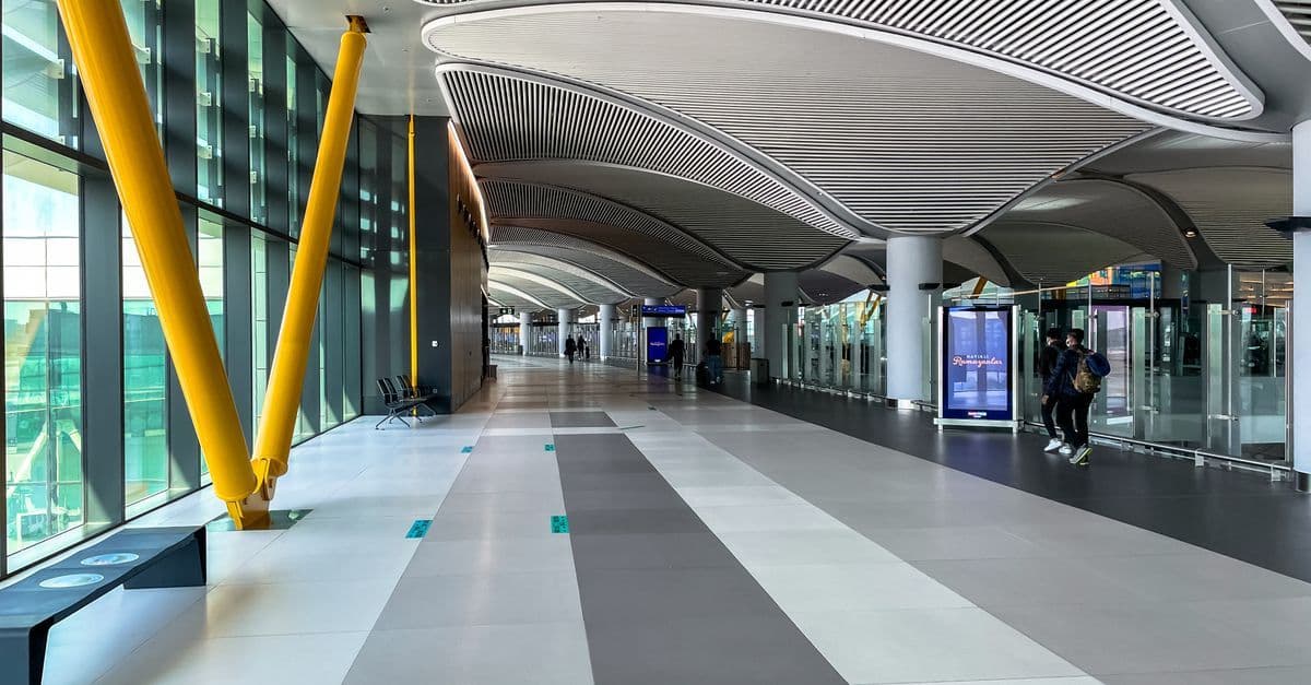 Reisende mit Rucksack in einer hellen Halle des Flughafens Istanbul mit gelben Stützen und großer Glasfront.