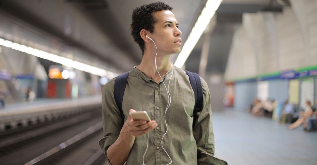 Ein Reisender mit Rucksack und Kopfhörern hält am Bahnsteig sein Smartphone in der Hand.