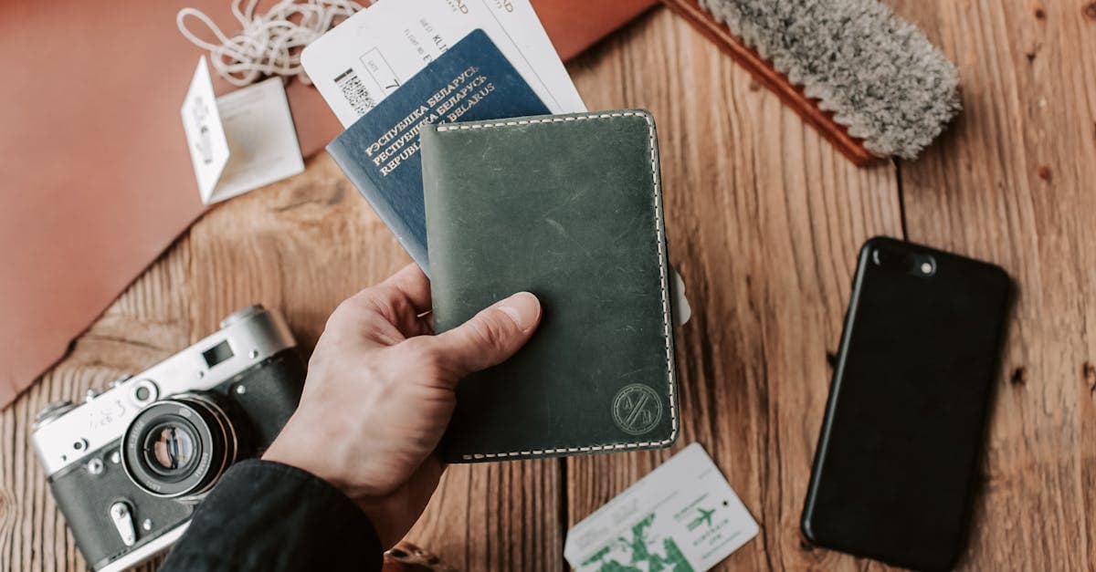 Mano sosteniendo pasaporte junto a cámara, teléfono y boletos sobre mesa de madera para solicitud de documentos.