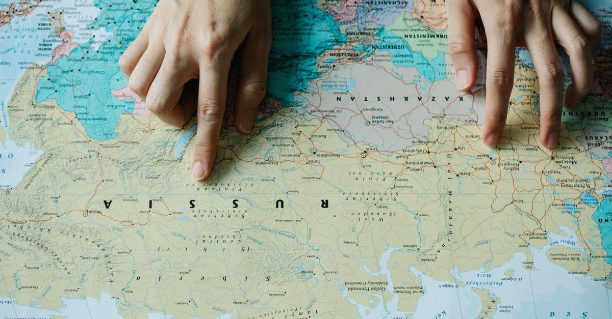 Mãos apontando para a Rússia em mapa-múndi detalhado, ilustrando a localização dos overseas countries and territories.