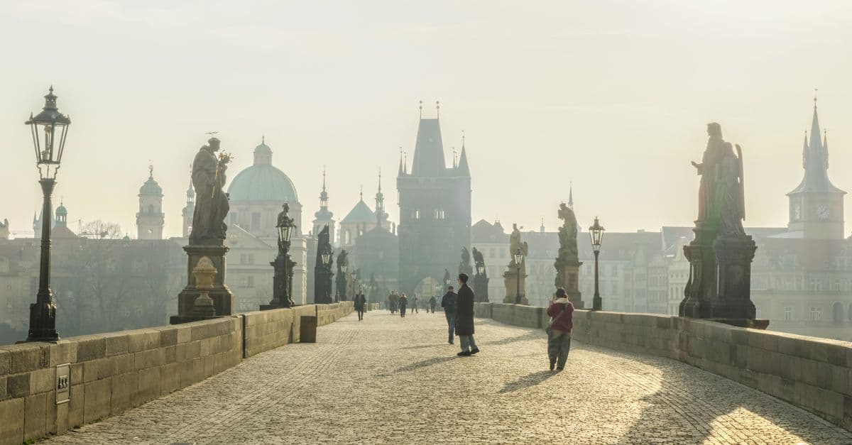 People walk across Prague’s Charles Bridge toward the city’s historic skyline in the morning haze.