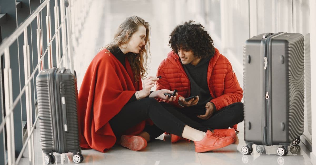 Two travelers sit among their suitcases, looking at a phone while planning their trip.