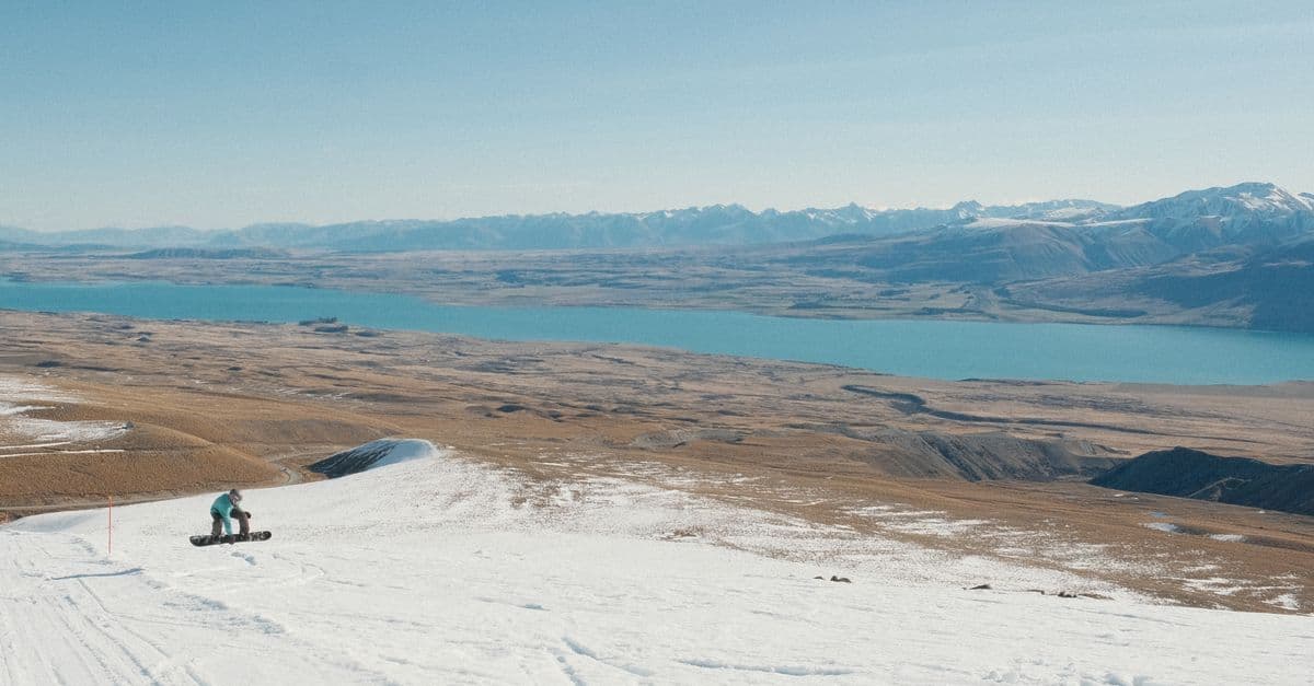 A lone snowboarder rides a groomed slope above Lake Tekapo in New Zealand, with snowy mountains behind.