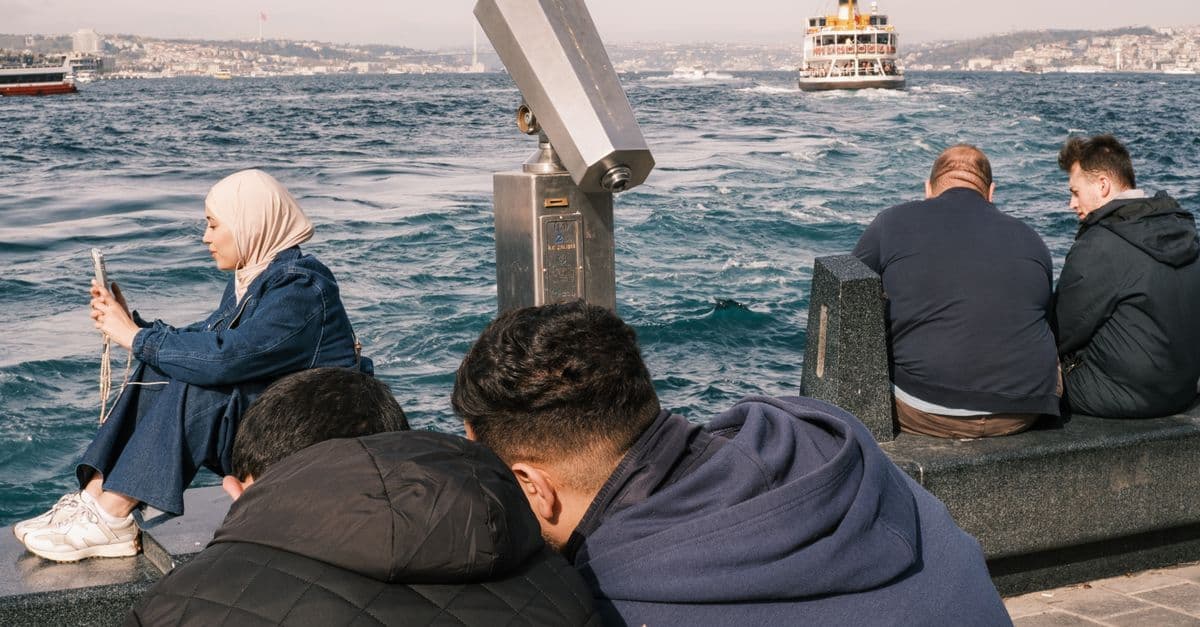 Eine Frau schaut am Bosporus in Istanbul auf ihr Handy, im Hintergrund fährt eine Fähre.