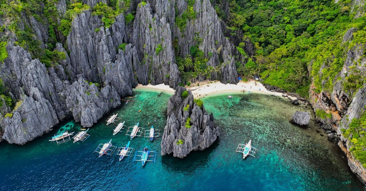 Türkisblaues Wasser, Kalksteinfelsen und Boote umrahmen einen kleinen Sandstrand auf den Philippinen.