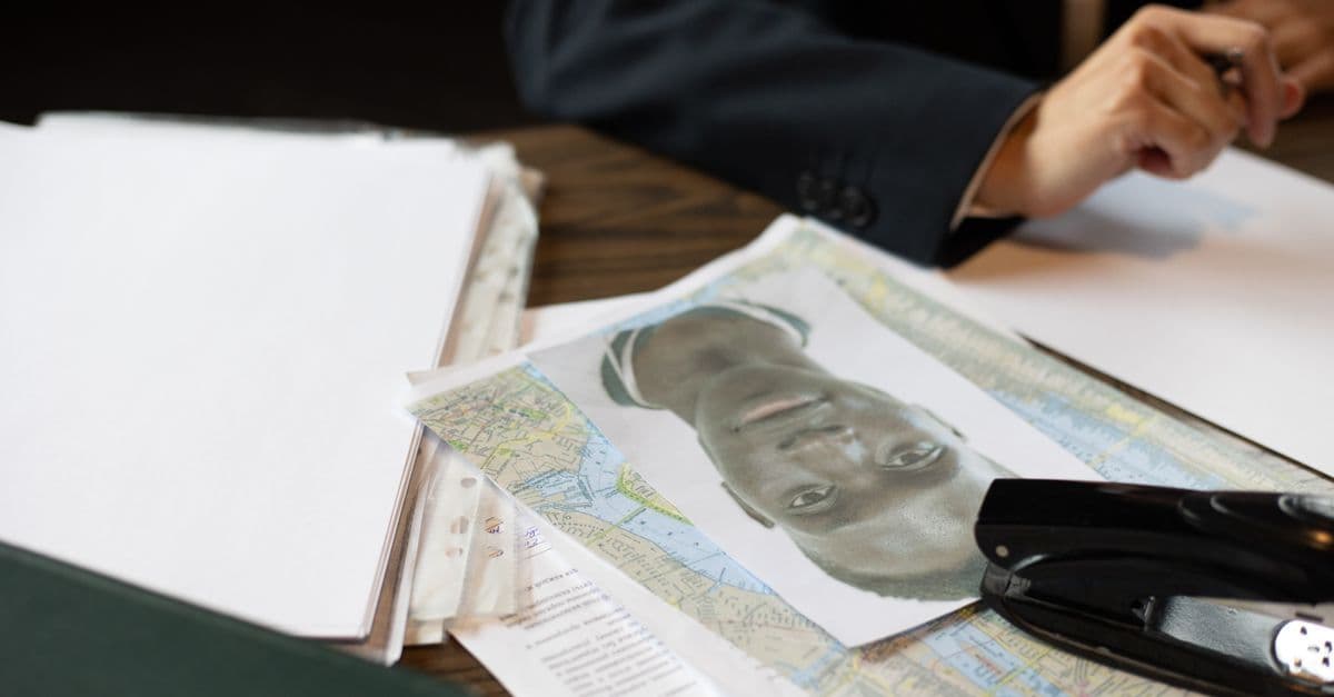 A printed portrait photo lies on a map and paperwork beside a stapler, as a hand holds a pen nearby.