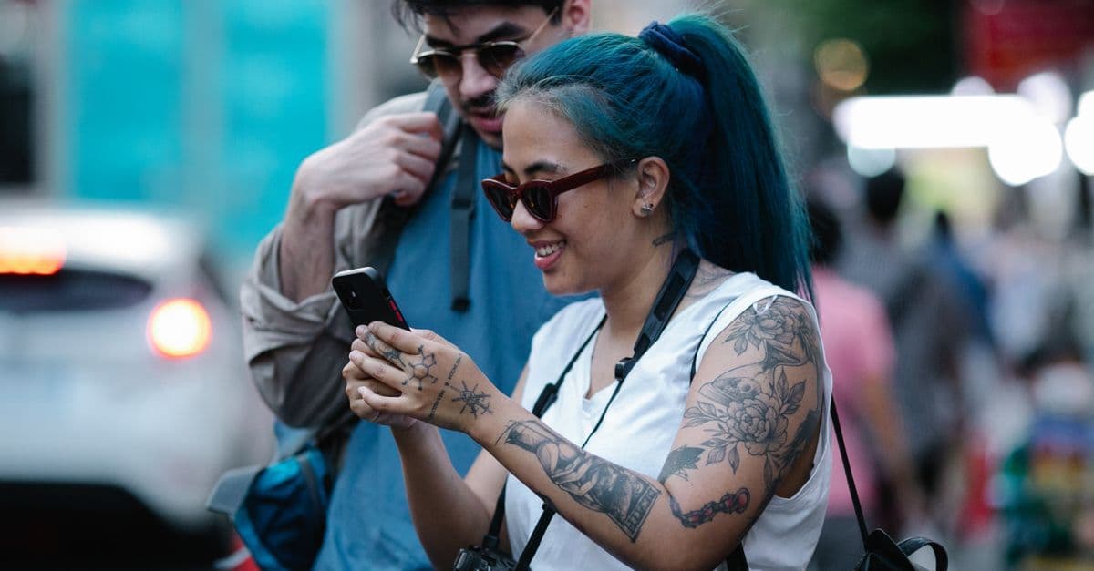 Two travelers with backpacks smile at a phone together on a busy city street.