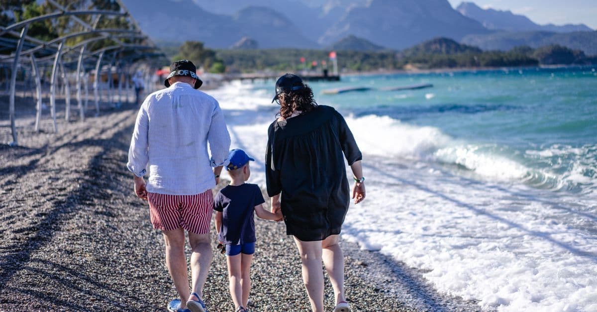 Rodzina z dzieckiem idzie kamienistą plażą w Kemerze wzdłuż turkusowego morza, w tle góry.