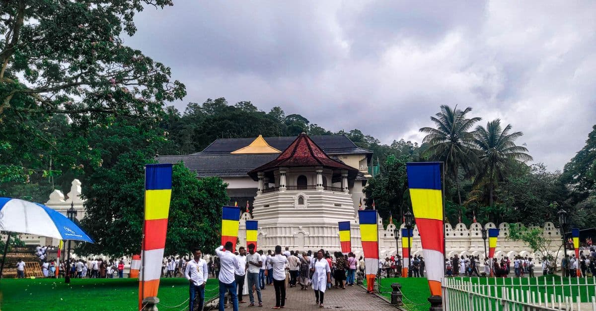 Visitors gather outside a white temple with a tiered red roof and colorful striped pillars in a green courtyard.