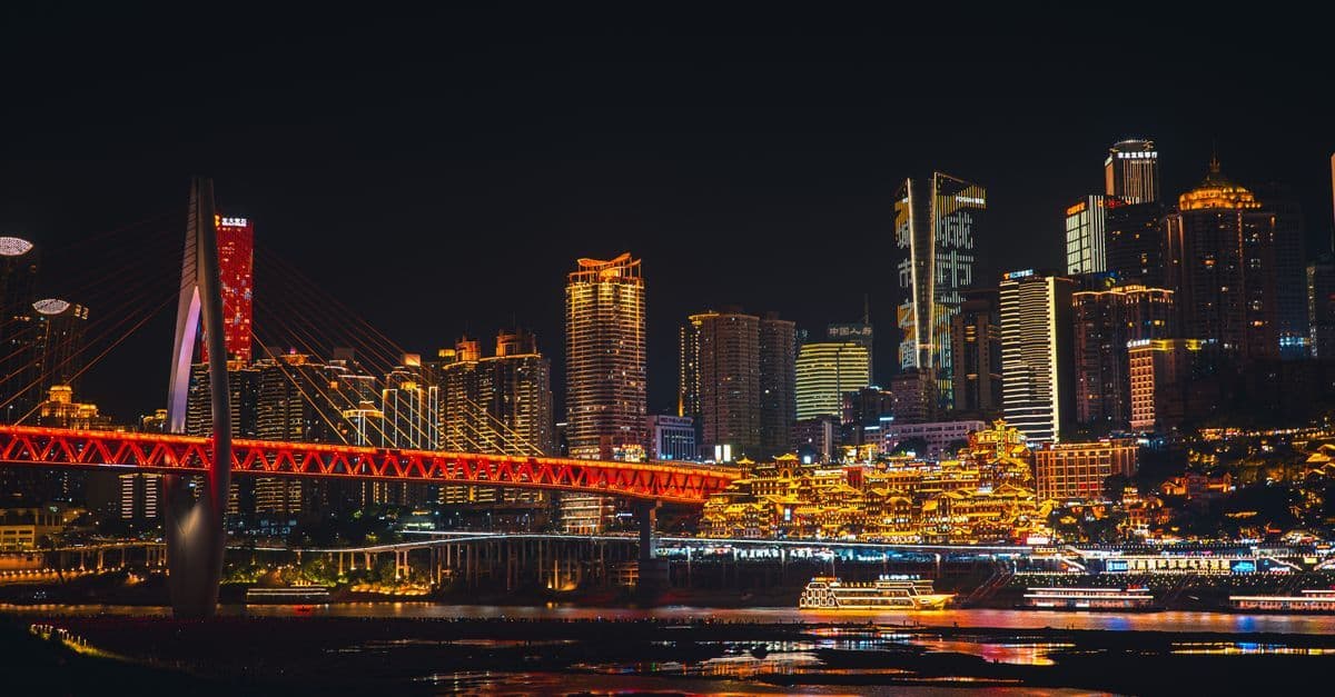 A brightly lit bridge, skyscrapers, and boats line the river in Chongqing at night.