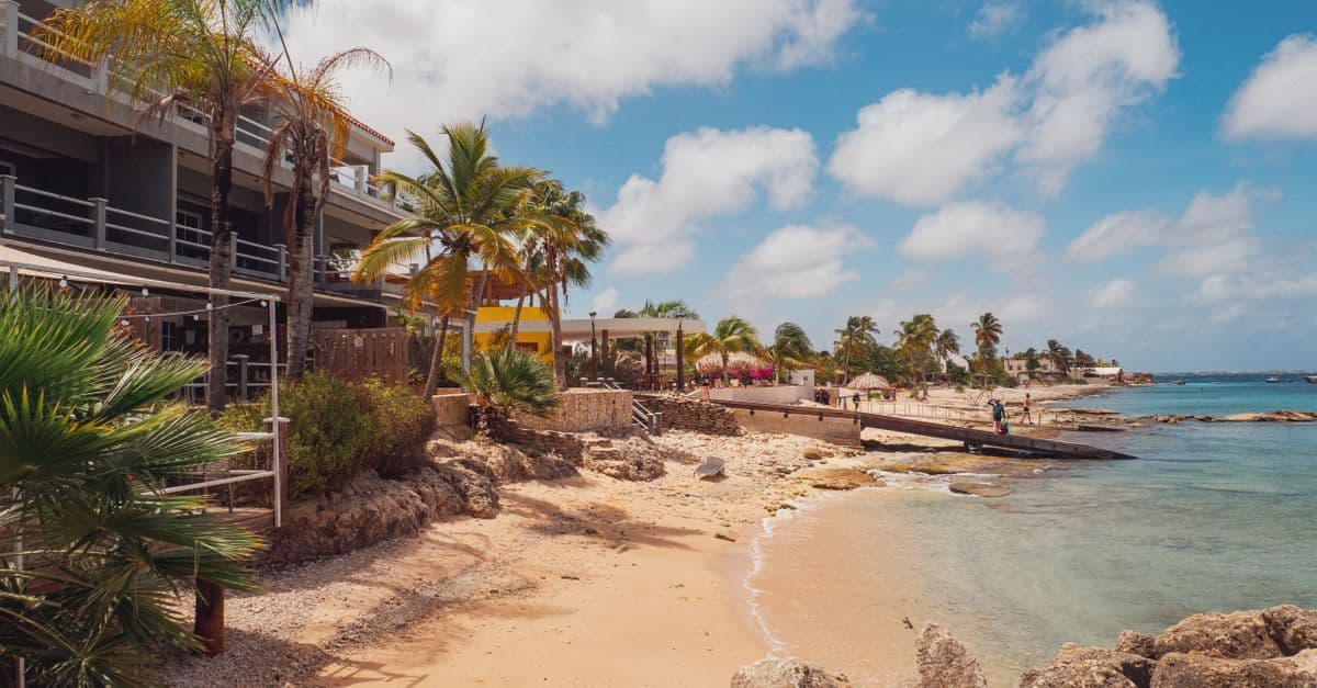 A sandy Caribbean shoreline beside resort buildings and palm trees, with calm turquoise water and a few people.
