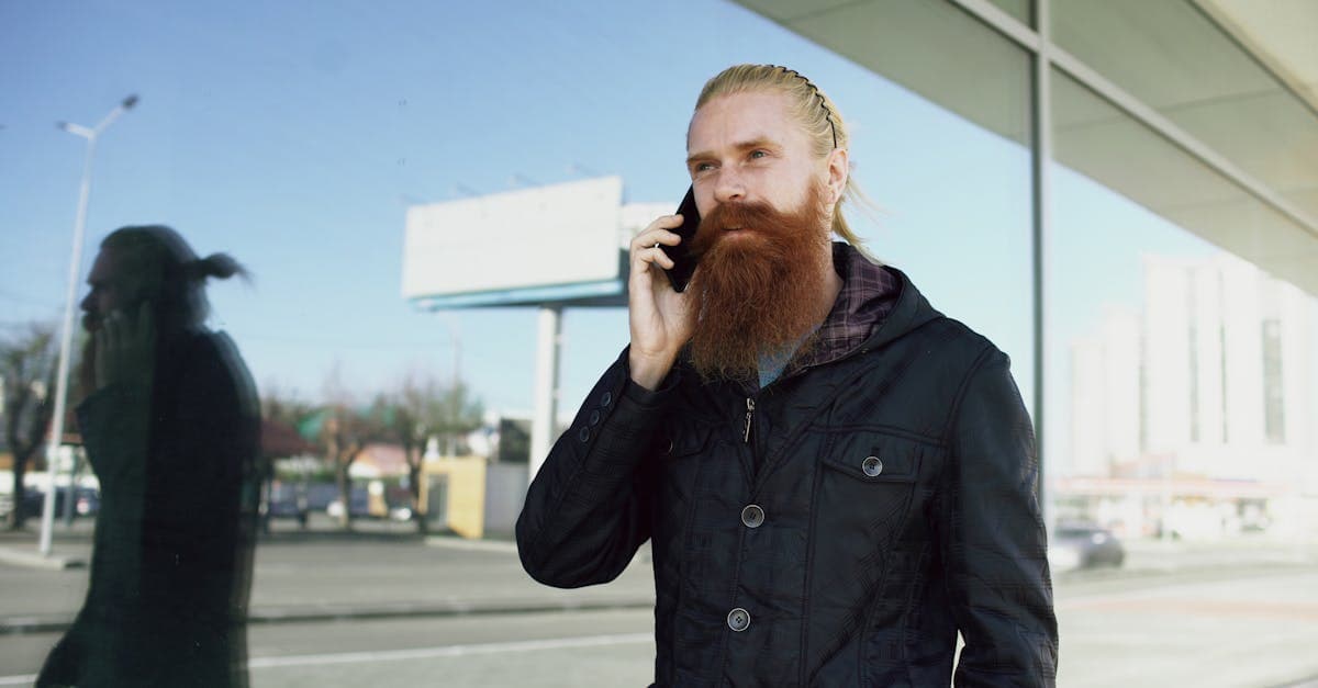 A man with a long red beard using a smartphone outside a glass building on a sunny day.