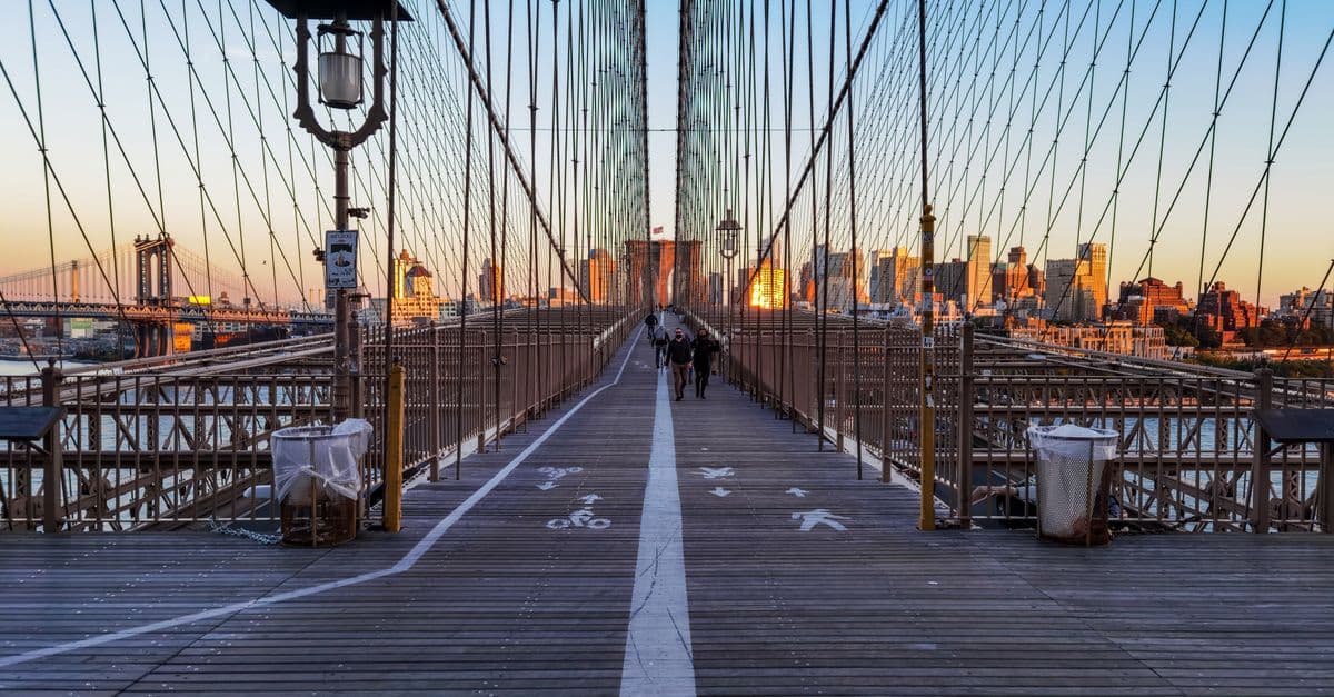 Pedoni sulla passerella in legno del ponte di Brooklyn al tramonto, tra i cavi d'acciaio e i palazzi di New York.