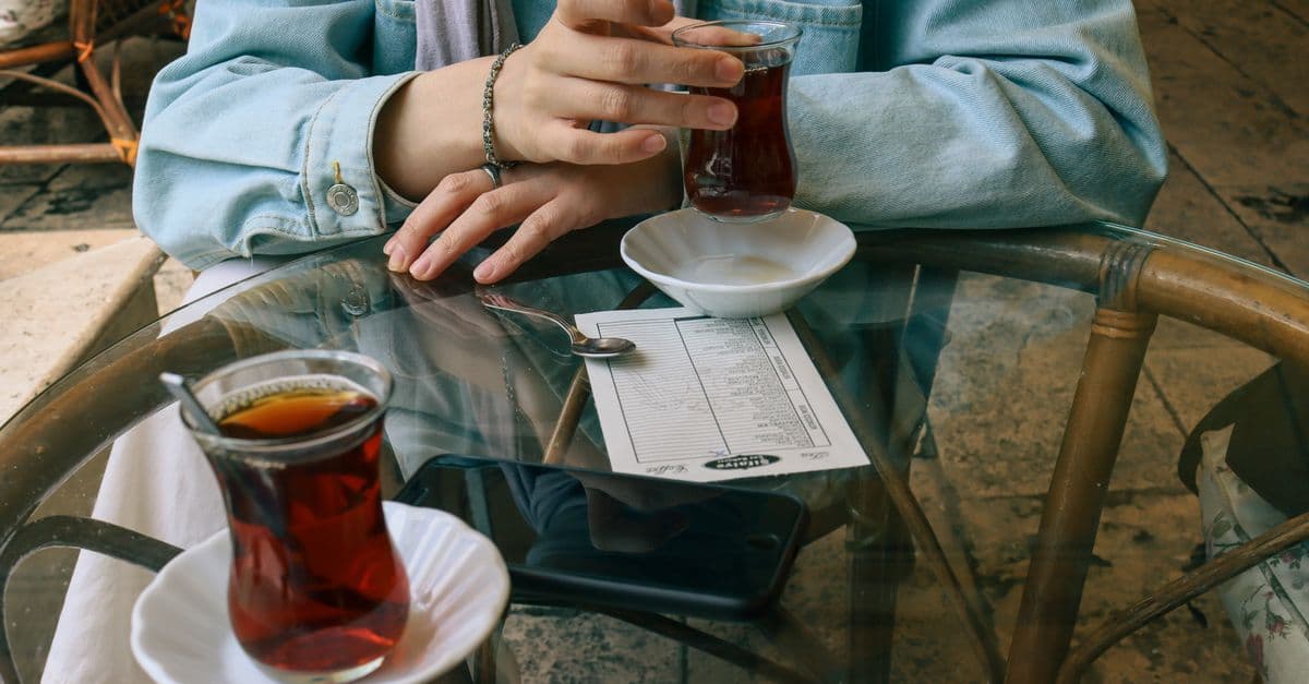 Eine Person hält ein Glas Tee am Glastisch, auf dem ein Smartphone, ein Zettel und ein zweites Teeglas liegen.