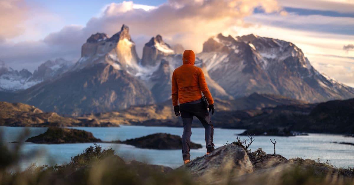 A hiker in an orange jacket looks across a Patagonian lake toward rugged mountains at sunset.