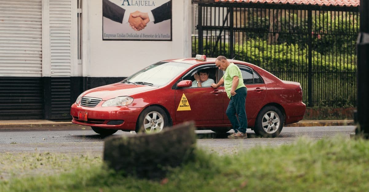 A man speaks with passengers through the window of a red taxi parked on a street in Alajuela.