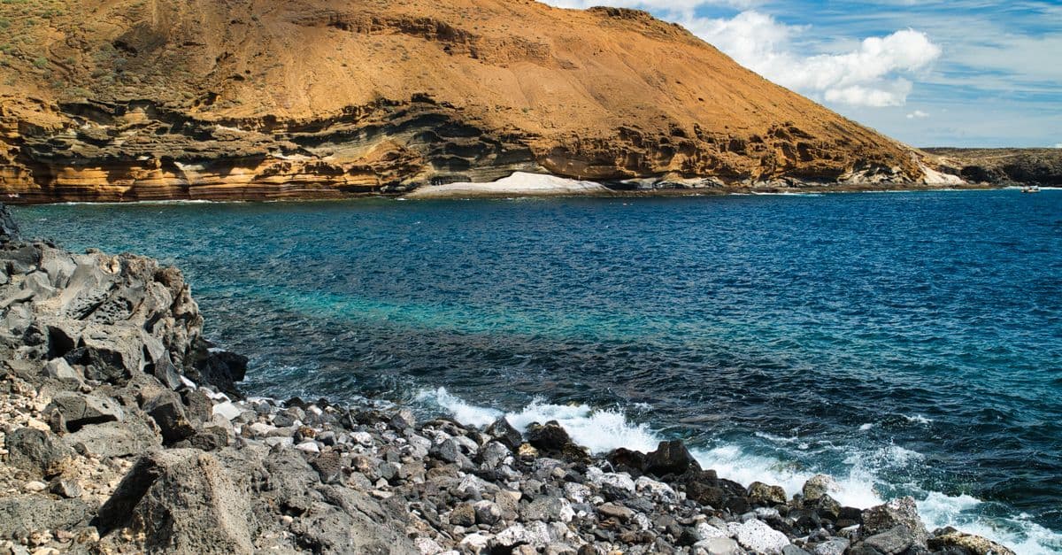 Scogliera rocciosa e mare turchese ai piedi di aride montagne vulcaniche, sotto un cielo parzialmente nuvoloso.