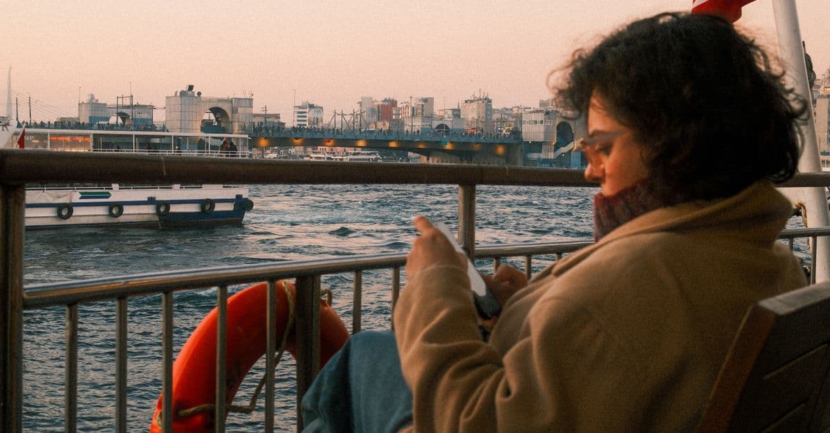 Eine Frau blickt auf einer Fähre in Istanbul auf ihr Smartphone, dahinter liegen Schiffe und die Stadt am Wasser.