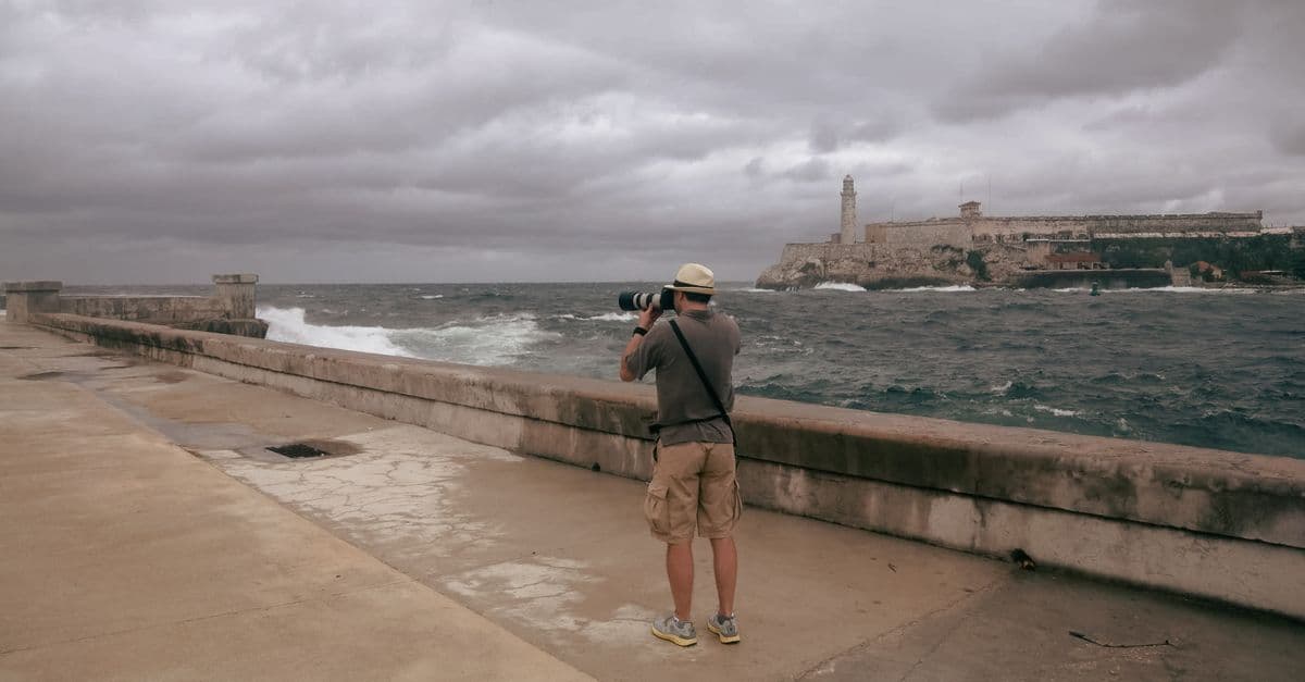 Un uomo fotografa il mare mosso e il cielo coperto dal lungomare dell’Avana, vicino a una fortezza.
