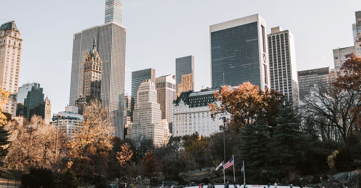 Skaters gather on a Central Park ice rink beneath Manhattan skyscrapers and autumn-colored trees.
