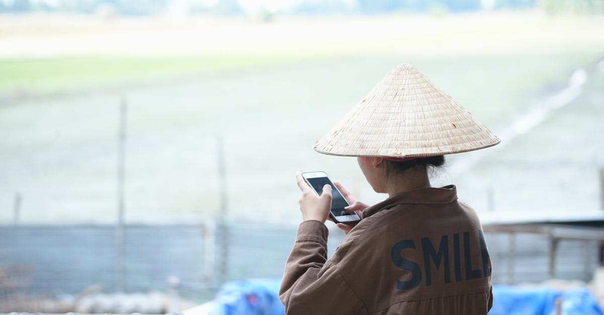 Back view of an Asian woman wearing a conical hat using a smartphone outdoors.