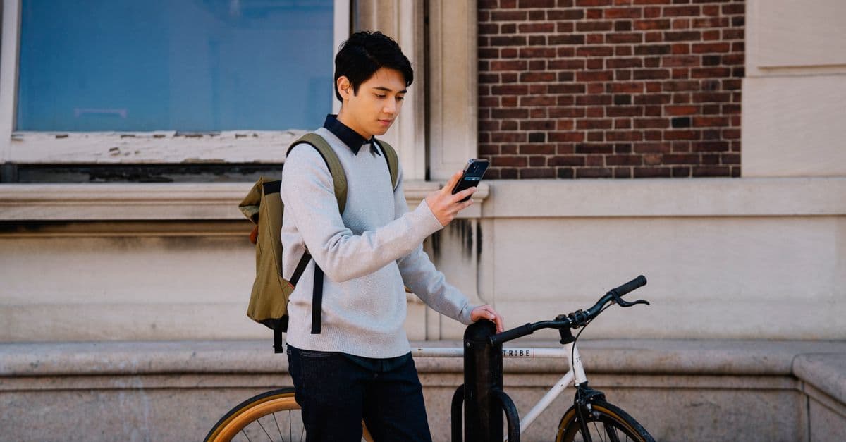 Joven con mochila apoyado en su bicicleta mirando el celular frente a un edificio urbano.
