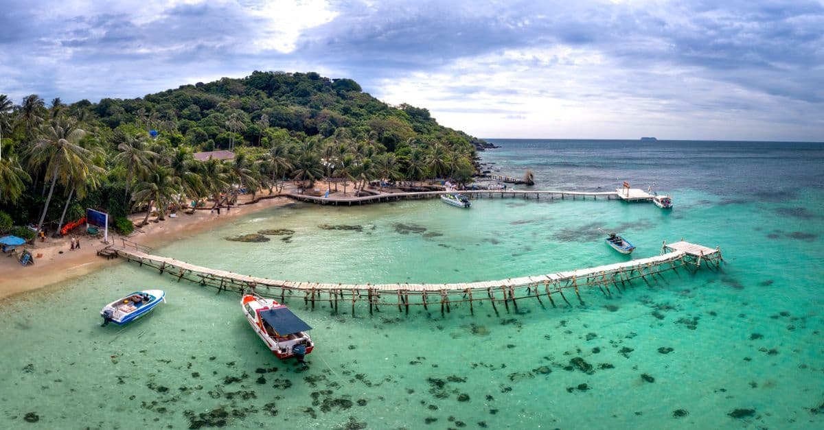 A palm-lined Belize cay is surrounded by turquoise water, with boats moored beside long wooden piers.