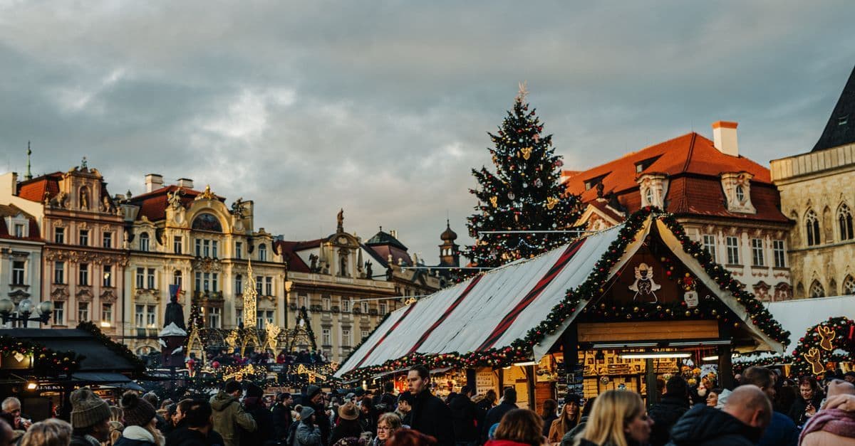 Crowds gather around a decorated Christmas tree and wooden market stalls in a historic European square.