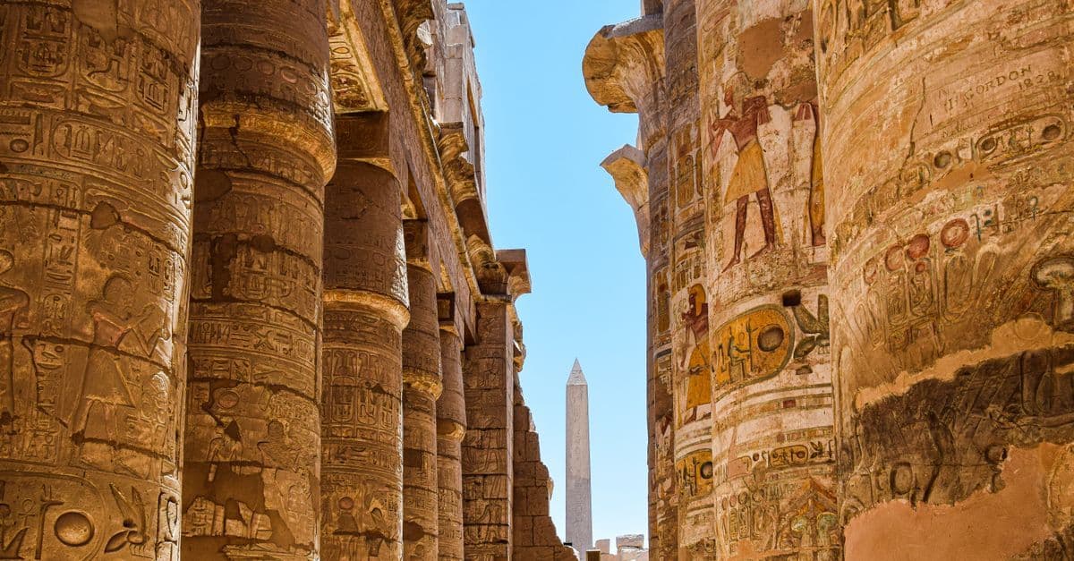 Columnas de un templo egipcio con relieves y un obelisco al fondo, bajo un cielo despejado.