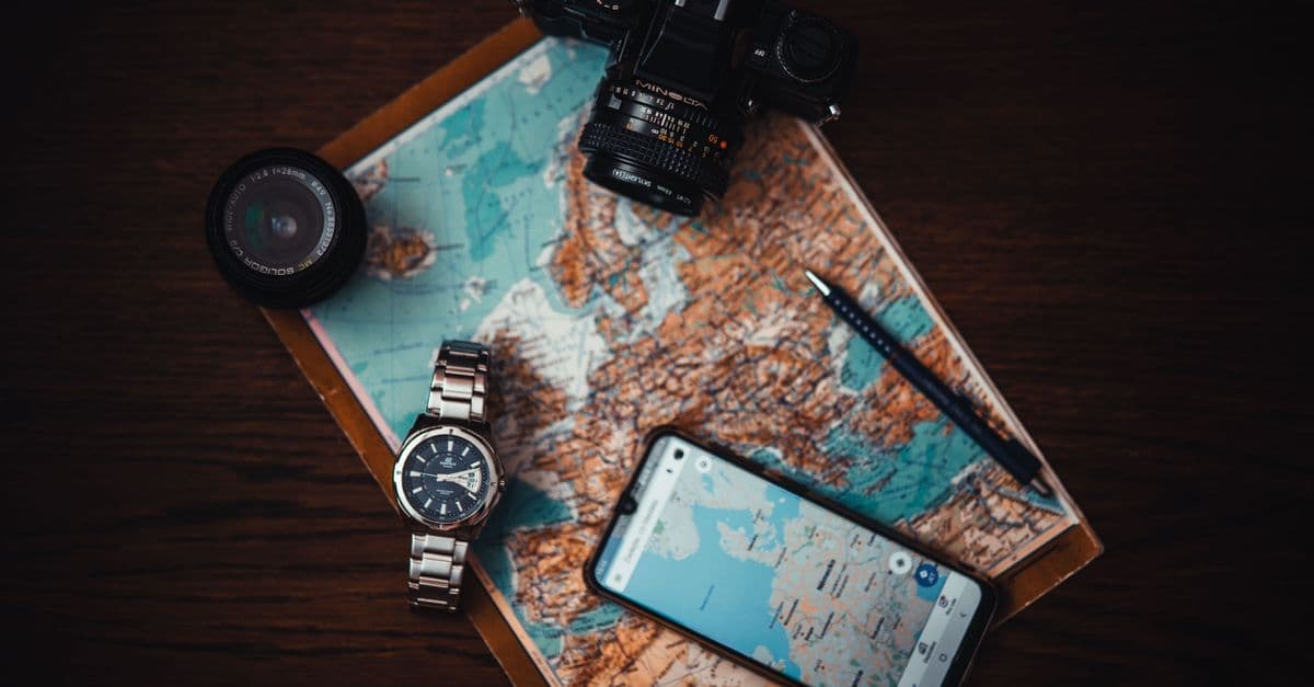 Accessori da viaggio essenziali visti dall'alto: mappa, fotocamera, smartphone e orologio su superficie in legno.