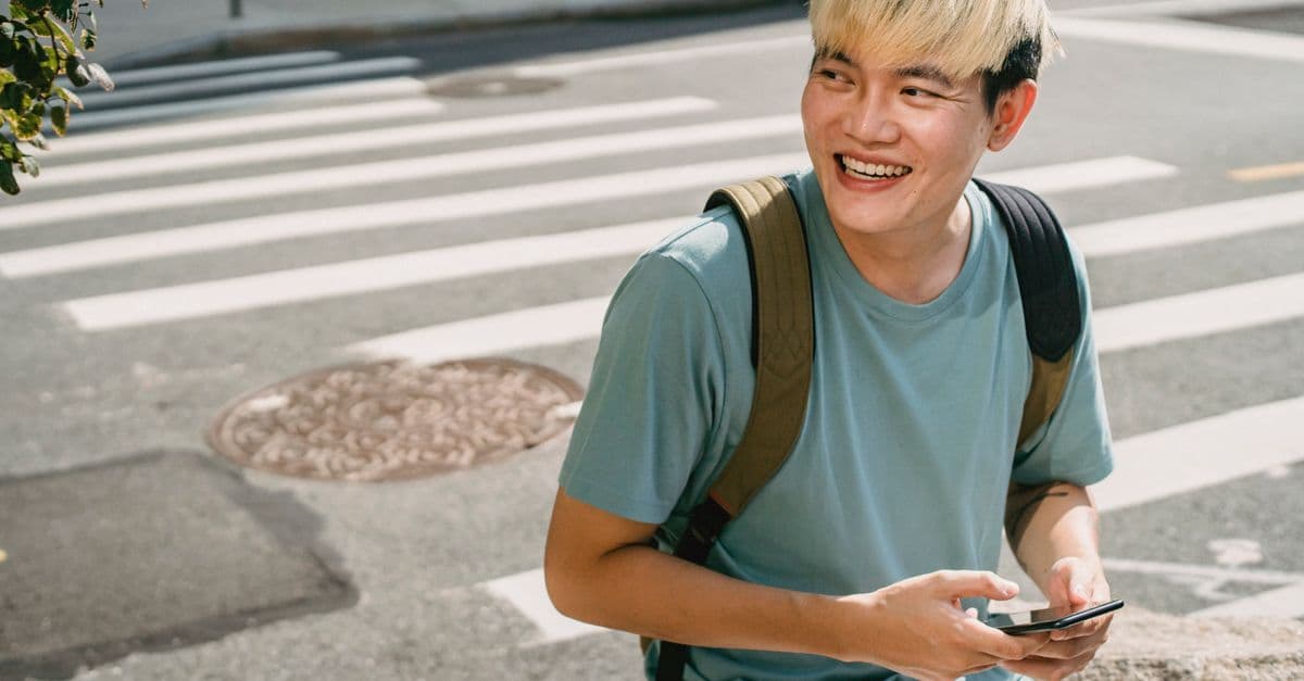 Un ragazzo con lo zaino sorride con lo smartphone in mano, accanto alle strisce pedonali di una strada.