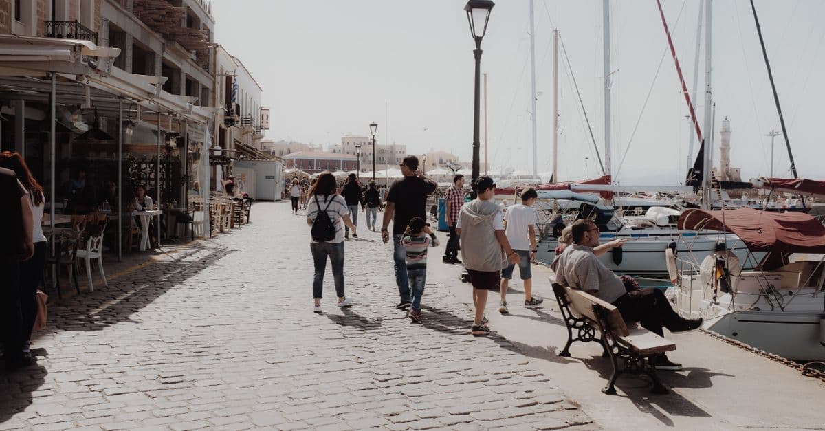 Familien spazieren an Cafés und festgemachten Segelbooten entlang einer sonnigen Hafenpromenade.