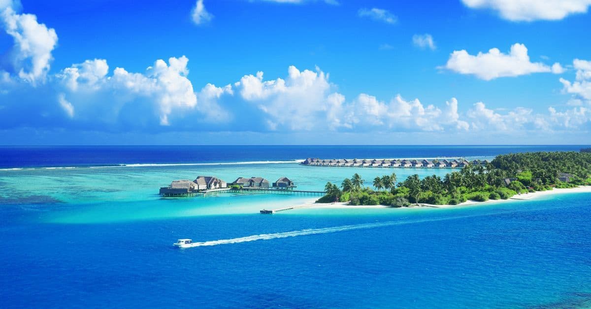 A sun-drenched Maldives resort with crystal-clear waters beneath a brilliant blue sky at peak season.