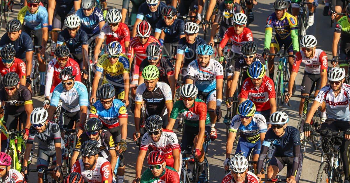 Gruppo di ciclisti in maglia colorata in gara su strade urbane durante una tappa di ciclismo professionistico.