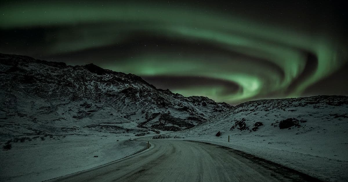 Una carretera nevada atraviesa montañas bajo una intensa aurora boreal en el cielo nocturno de Islandia.