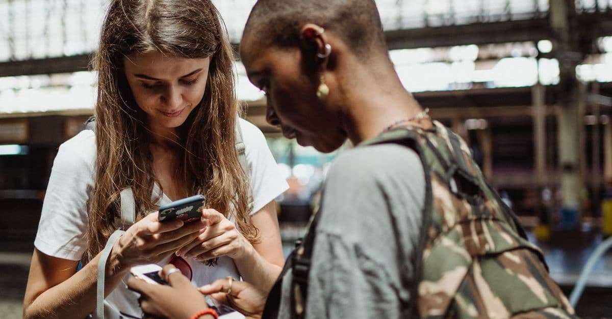 Zwei Reisende mit Rucksäcken schauen in einer Bahnhofshalle auf ihre Smartphones.