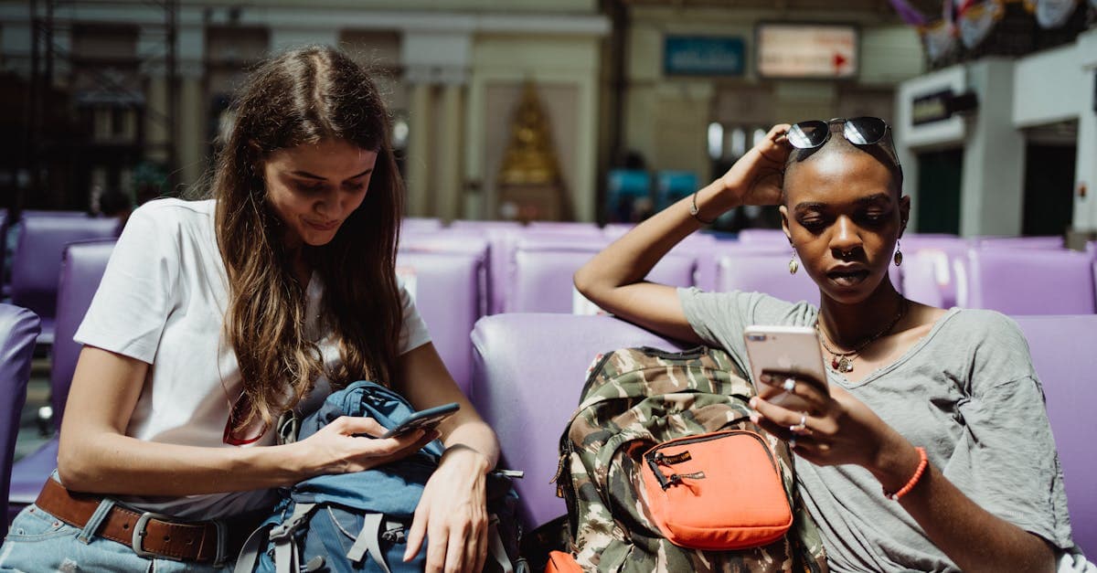 Two travelers using smartphones at a railway station, staying connected to play Gartic Phone while traveling